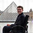 Portrait souriant de Pierre, un homme en fauteuil roulant, devant la pyramide du Louvre.